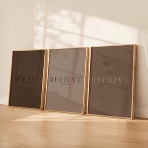 May include: Three framed art prints with inspirational words. The prints feature the words "PRAY", "BELIEVE", and "RECEIVE" in a serif font. The frames are a light wood colour, and the background is a light beige. The overall colour palette is neutral.