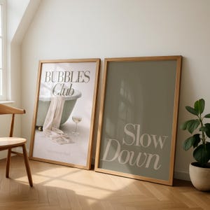 Peut inclure: Deux affiches encadrées avec des cadres en bois. L'une présente une baignoire avec le texte "Bubbles Club" et "an evening ritual". L'autre affiche affiche les mots "Slow Down" en clair sur un fond vert.
