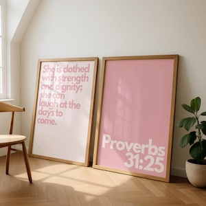 May include: Two framed art prints with wooden frames. One print has a white background with pink text that reads "She is clothed with strength and dignity; she can laugh at the days to come." The other print has a pink background with white text that reads "Proverbs 31:25."