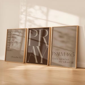 May include: Three framed art prints with gold-coloured frames. The prints feature inspirational quotes and Bible verses in a neutral colour palette. The middle print says "PRAY" in large letters. The other prints have text from Proverbs 31:25 and Psalm 46:5.