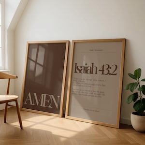 May include: Two framed art prints with wooden frames. One print is brown with the word "AMEN" in large white letters. The other print is beige with the text "Isaiah 43:2" and a Bible verse. A plant in a pot is next to the prints.