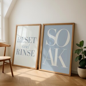 May include: Two framed art prints with wooden frames. One print has the words "RESET and RINSE" in blue on a white background. The other print has the word "SOAK" in white on a light blue background. A wooden chair and a potted plant are also visible.