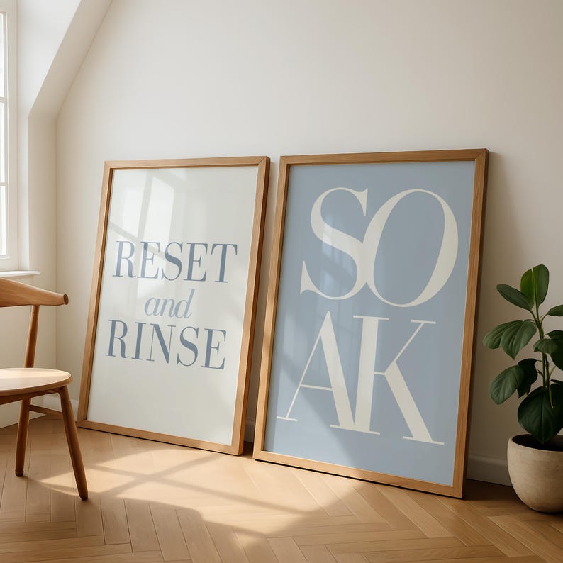 May include: Two framed art prints with wooden frames. One print has the words "RESET and RINSE" in blue on a white background. The other print has the word "SOAK" in white on a light blue background. A wooden chair and a potted plant are also visible.