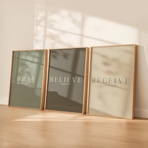 May include: Three framed art prints with inspirational text. The prints are in a row, each with a different colour background: green, grey, and beige. The text on the prints reads "PRAY", "BELIEVE", and "RECEIVE". The frames are a light wood colour.