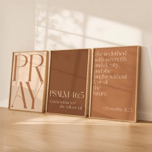 以下が含まれることがあります： インスピレーションを与える引用句が書かれた3つの額入りアートプリント。最初のプリントには大きな文字で「PRAY」と書かれています。2番目のプリントには「PSALM 46:5」と引用句が書かれています。3番目のプリントには箴言31:25からの引用句が書かれています。すべてのプリントは茶色とベージュの色使いです。