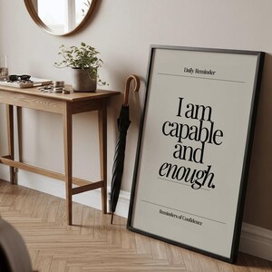 May include: A framed print with the text "I am capable and enough" in a black frame. A black umbrella with a wooden handle leans against the wall. A wooden table with a plant and accessories is in the background.