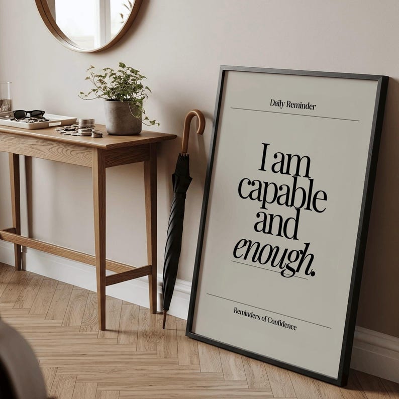 May include: A framed print with the text "I am capable and enough" in a black frame. A black umbrella with a wooden handle leans against the wall. A wooden table with a plant and accessories is in the background.