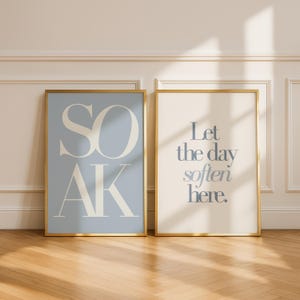 May include: Two framed art prints with gold frames. One print has the word "SOAK" in large, stylized letters on a light blue background. The other print says "Let the day soften here" on a cream background.