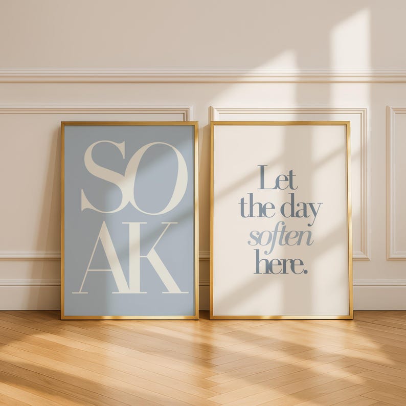May include: Two framed art prints with gold frames. One print has the word "SOAK" in large, stylized letters on a light blue background. The other print says "Let the day soften here" on a cream background.