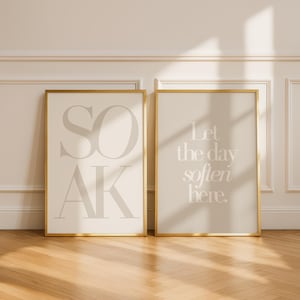 May include: Two framed art prints with gold frames. The left print says "SOAK" in large, light gray letters on a cream background. The right print says "Let the day soften here" in light gray on a beige background. The prints are displayed on a wooden floor.