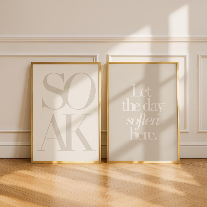 May include: Two framed art prints with gold frames. The left print says "SOAK" in large, light gray letters on a cream background. The right print says "Let the day soften here" in light gray on a beige background. The prints are displayed on a wooden floor.