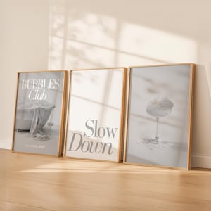 Könnte beinhalten: Drei gerahmte Kunstdrucke mit Holzrahmen. Der linke Druck zeigt "Bubbles Club" mit einem Badewannenbild. Der mittlere Druck zeigt "Slow Down". Der rechte Druck zeigt ein Cocktailglas. Alle Drucke haben eine neutrale Farbpalette.
