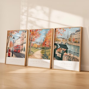 May include: Three framed vertical art prints featuring autumn city scenes. The left print shows a red bus in Fitzrovia, the center print depicts Central Park with fall foliage, and the right print displays the La Seine river in Paris.