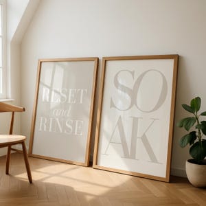 May include: Two framed art prints with light beige backgrounds and wooden frames. One print reads "RESET and RINSE" in white text, the other reads "SOAK" in large, stylized letters. A wooden chair and potted plant are also visible.