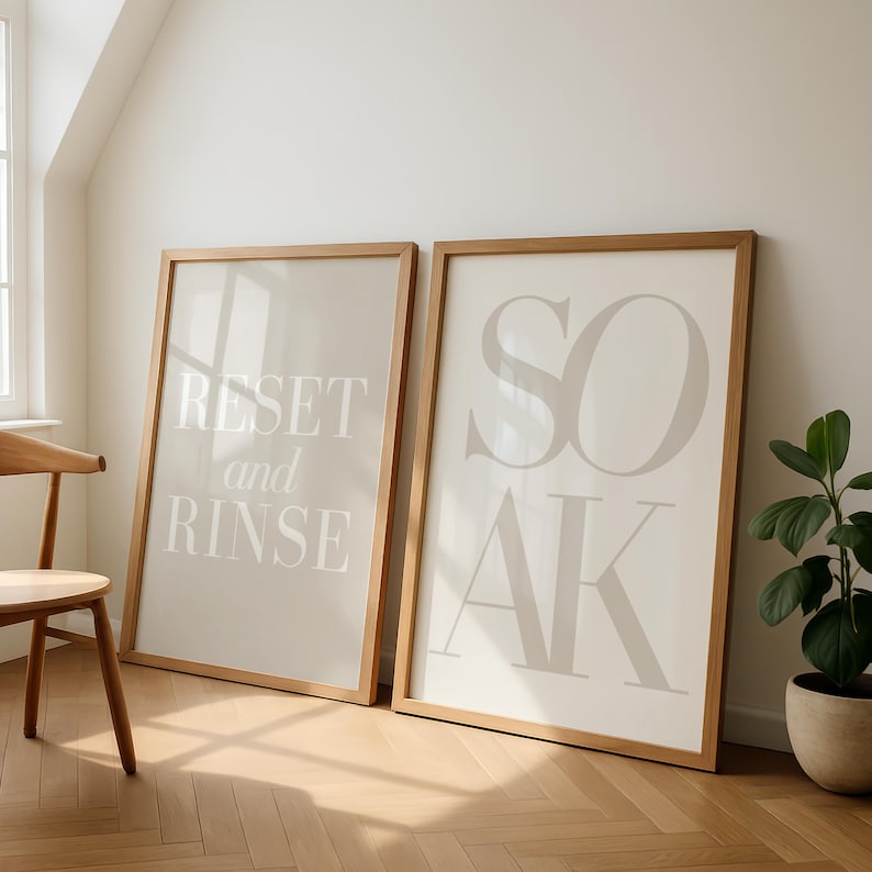 May include: Two framed art prints with light beige backgrounds and wooden frames. One print reads "RESET and RINSE" in white text, the other reads "SOAK" in large, stylized letters. A wooden chair and potted plant are also visible.
