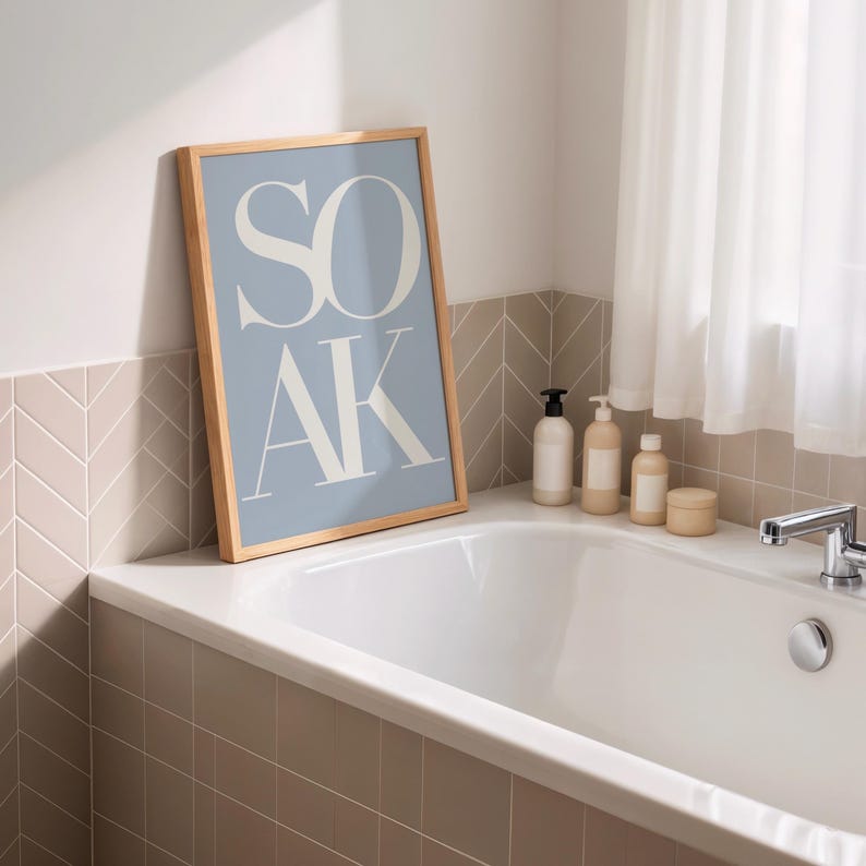 May include: A framed poster with the word "SOAK" in large white letters on a light blue background. The poster is in a wooden frame and is leaning against a bathroom wall. The bathroom has a white bathtub and several bottles of bath products.