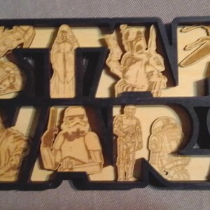 May include: Wooden Star Wars sign with the Star Wars logo in black, filled with laser-cut images of characters and vehicles from the Star Wars franchise. The sign is made of light-colored wood.