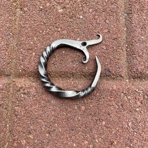 May include: A hand-forged metal bottle opener with a twisted handle and a curved hook. The opener is silver in color and sits on a red brick surface. The design is simple and rustic.