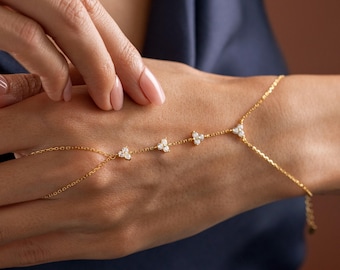 Op de afbeelding: Een delicate gouden handketting met kleine, sprankelende, heldere edelstenen in een bloemmotief. De ketting verbindt met een armband en wikkelt zich om de vingers. De sieraden worden gepresenteerd tegen een donkerblauwe achtergrond.
