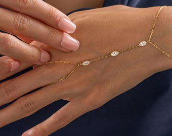 Op de afbeelding: Een delicate gouden handkettingarmband met vier ovaalvormige, heldere edelstenen. De armband verbindt met een ring en wikkelt zich om de achterkant van de hand. De sieraden worden getoond op een hand met een donkerblauwe achtergrond.