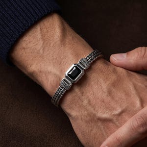 May include: A silver bracelet featuring a braided chain design and a central rectangular setting containing a polished black stone. The metal components display intricate engraved geometric patterns along the edges of the central focal piece.