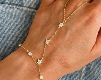 May include: A gold-colored hand chain bracelet featuring star and round-shaped embellishments. The bracelet connects from the wrist to the finger with delicate chain links. The stars and round embellishments are encrusted with small, clear stones.