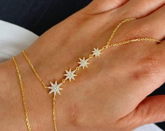 May include: A gold-colored hand chain with a bracelet and a ring chain, connected by a delicate chain. The chain features five star-shaped charms, each encrusted with small, sparkling crystals. The jewelry is worn on a hand.