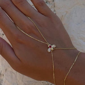 Puede incluir: Delicada cadena de mano dorada con un dije en forma de abeja con una gema roja en el centro y pequeñas piedras claras. La cadena se extiende a la muñeca y al dedo, creando un accesorio elegante. Joya exhibida sobre un fondo neutro.