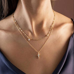 Op de afbeelding: Een gelaagde gouden ketting met drie strengen. De bovenste streng heeft kleine, ronde accenten en een bungelende heldere edelsteen. De middelste streng heeft een reeks kleine, platte accenten. De onderste streng heeft een stervormige hanger met een helder edelsteen centrum.