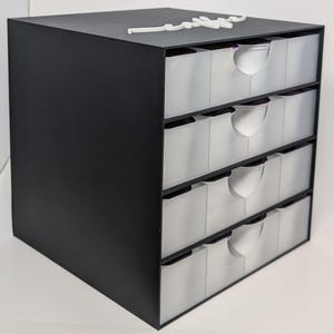Pode incluir: Uma caixa de armazenamento preta com quatro gavetas transl&uacute;cidas e puxadores brancos. Um elemento decorativo branco est&aacute; no topo. A caixa foi projetada para organizar itens pequenos.