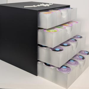Pode incluir: Uma unidade de armazenamento de c&aacute;psulas de caf&eacute; preta com quatro gavetas retr&aacute;teis. Cada gaveta &eacute; preenchida com c&aacute;psulas de caf&eacute; em v&aacute;rias cores. A unidade &eacute; feita de madeira preta e pl&aacute;stico transparente.