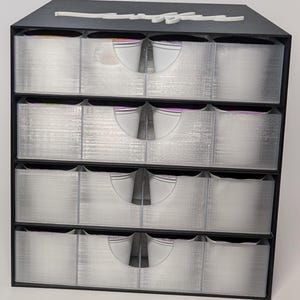 Pode incluir: Um organizador de armazenamento preto de quatro n&iacute;veis com gavetas transparentes e extra&iacute;veis. Cada gaveta tem uma al&ccedil;a branca curva. A parte superior do organizador tem um design decorativo branco em it&aacute;lico. As gavetas parecem estar vazias.