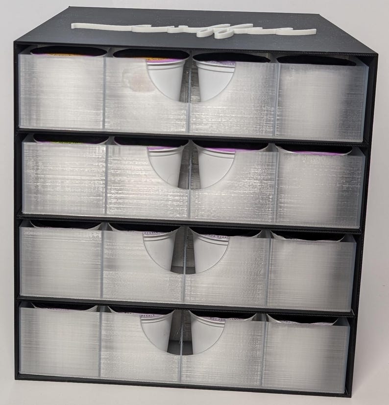 Pode incluir: Um organizador de armazenamento preto de quatro n&iacute;veis com gavetas transparentes e extra&iacute;veis. Cada gaveta tem uma al&ccedil;a branca curva. A parte superior do organizador tem um design decorativo branco em it&aacute;lico. As gavetas parecem estar vazias.