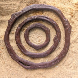 May include: Three concentric, dark brown, ring-shaped objects with a textured surface, arranged on a bed of sand. The rings vary in size, with the largest forming an outer spiral. The objects appear to be made of a natural material.