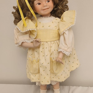 Puede incluir: Muñeca de porcelana con cabello rizado castaño y lazos amarillos. La muñeca viste un vestido amarillo con estampado floral, ribete de encaje blanco y zapatos negros. La muñeca está sobre un soporte transparente.