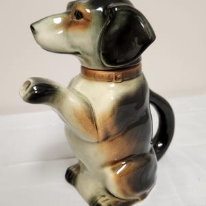 Może przedstawiać: Ceramiczny czajnik w kształcie psa z brązową obrożą. Czajnik jest głównie biały, z czarnymi, brązowymi i beżowymi akcentami. Pies siedzi wyprostowany z podniesioną łapą. Uchwyt jest czarny.