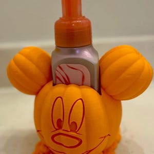 Puede incluir: Soporte para dispensador de jabón con forma de calabaza naranja con una cara sonriente y orejas grandes. El dispensador tiene una bomba y es naranja. El soporte está diseñado para sujetar una botella de jabón.