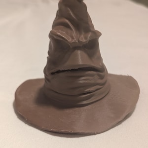 May include: A brown Sorting Hat figurine with a wide brim and a pointed top. The hat has a textured surface and a face-like appearance, with a mouth and furrowed brow. The hat is a replica of the Sorting Hat from the Harry Potter series.