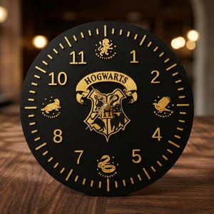 May include: Black round clock with gold-colored numerals and accents. The clock face features the word "HOGWARTS" and the Hogwarts crest. The clock also has the emblems of the four Hogwarts houses. The clock is on a wooden surface.