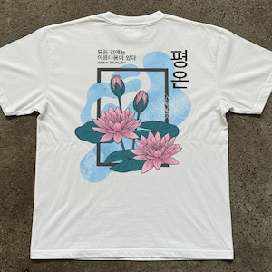 May include: White t-shirt featuring a floral design with pink lotus flowers, green leaves, and blue accents. The shirt has text in Korean. A black rectangle frames the flowers. The shirt is made of a soft material.