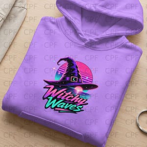May include: A light purple hooded sweatshirt featuring a graphic print of a dark witch hat set against a vibrant pink and blue sunset. The text "Witchy Waves" is printed in a stylized font below the hat, incorporating shades of pink and teal.
