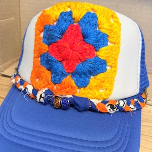 May include: A blue and white trucker hat featuring a square crochet patch on the front panel. The patch displays a red center surrounded by blue and orange yarn. A braided fabric trim in blue, orange, and white accents the brim with two beads.