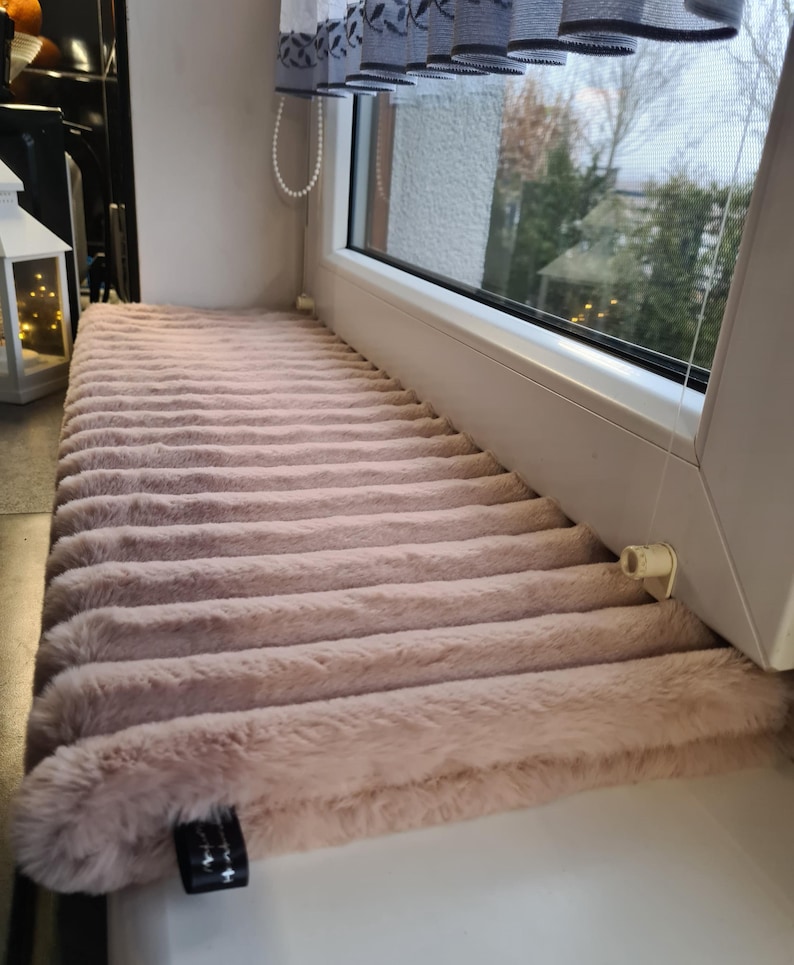 May include: A plush, light brown window seat cushion with a ribbed texture. The cushion is rectangular and fits snugly on a white windowsill. The soft, faux fur material adds a touch of warmth and comfort to the window area.