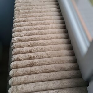 May include: A beige, corduroy-textured cat bed or window perch. The plush, rectangular cushion is made of soft, ridged fabric. The item is positioned near a window, suggesting a cozy spot for a pet to relax and observe.