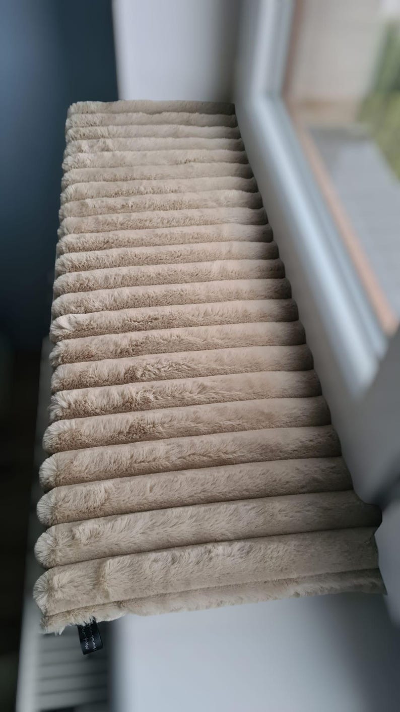 May include: A beige, corduroy-textured cat bed or window perch. The plush, rectangular cushion is made of soft, ridged fabric. The item is positioned near a window, suggesting a cozy spot for a pet to relax and observe.