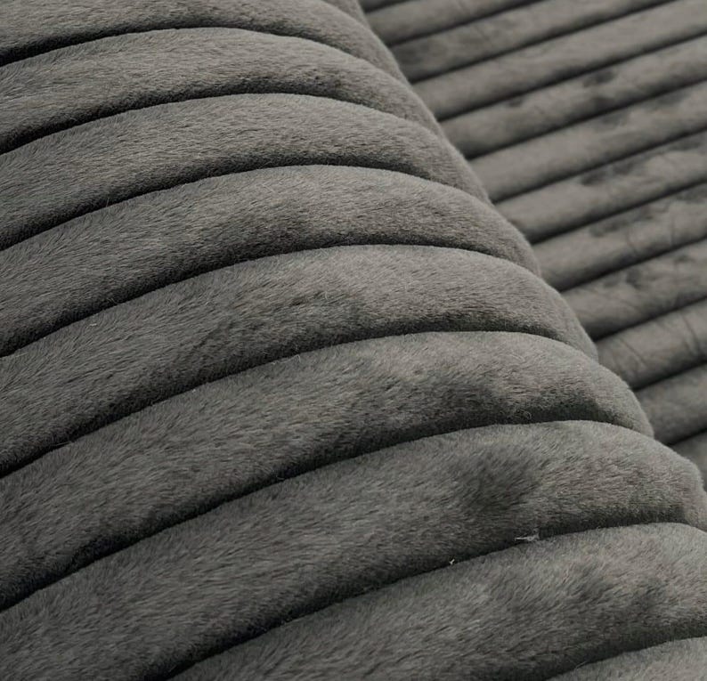 May include: Close-up of a gray, corduroy fabric with a ribbed texture. The fabric has a soft, plush appearance and is folded over, revealing the texture and depth of the material. The color is a muted, neutral gray.