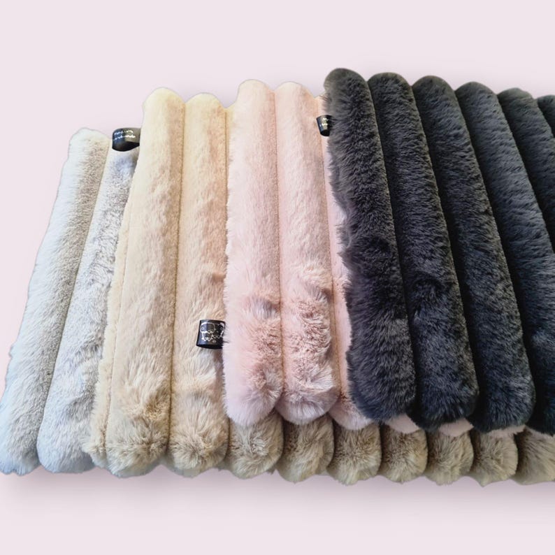 May include: A collection of plush, faux fur fabric in various colors, including white, beige, pink, and shades of gray. The fabric is arranged in vertical, ribbed sections, with a small tag attached to some pieces.