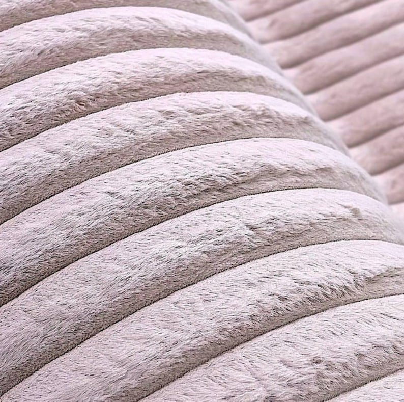 May include: Close-up of a soft, textured fabric in a light gray color. The fabric has a corduroy-like pattern with raised, rounded ridges running horizontally across the frame. The material appears plush and inviting, suggesting comfort and warmth.