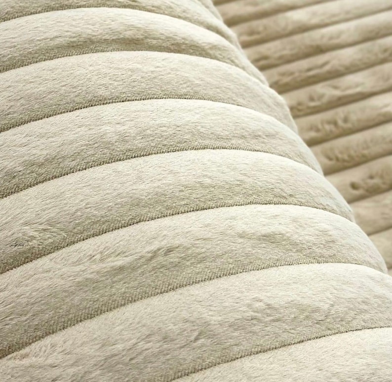 May include: Close-up of a soft, textured fabric in a light beige color. The fabric has a ribbed design, with parallel, rounded ridges creating a plush, inviting surface. The material appears to be a cozy, possibly plush, textile.
