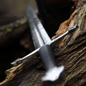 Pode incluir: Um close-up de uma espada prateada com uma empunhadura preta, repousando sobre uma superfície de madeira envelhecida. A lâmina da espada está angulada para o espectador, mostrando sua borda afiada e design intrincado. O texto "ForgedbyAlan" é visível.
